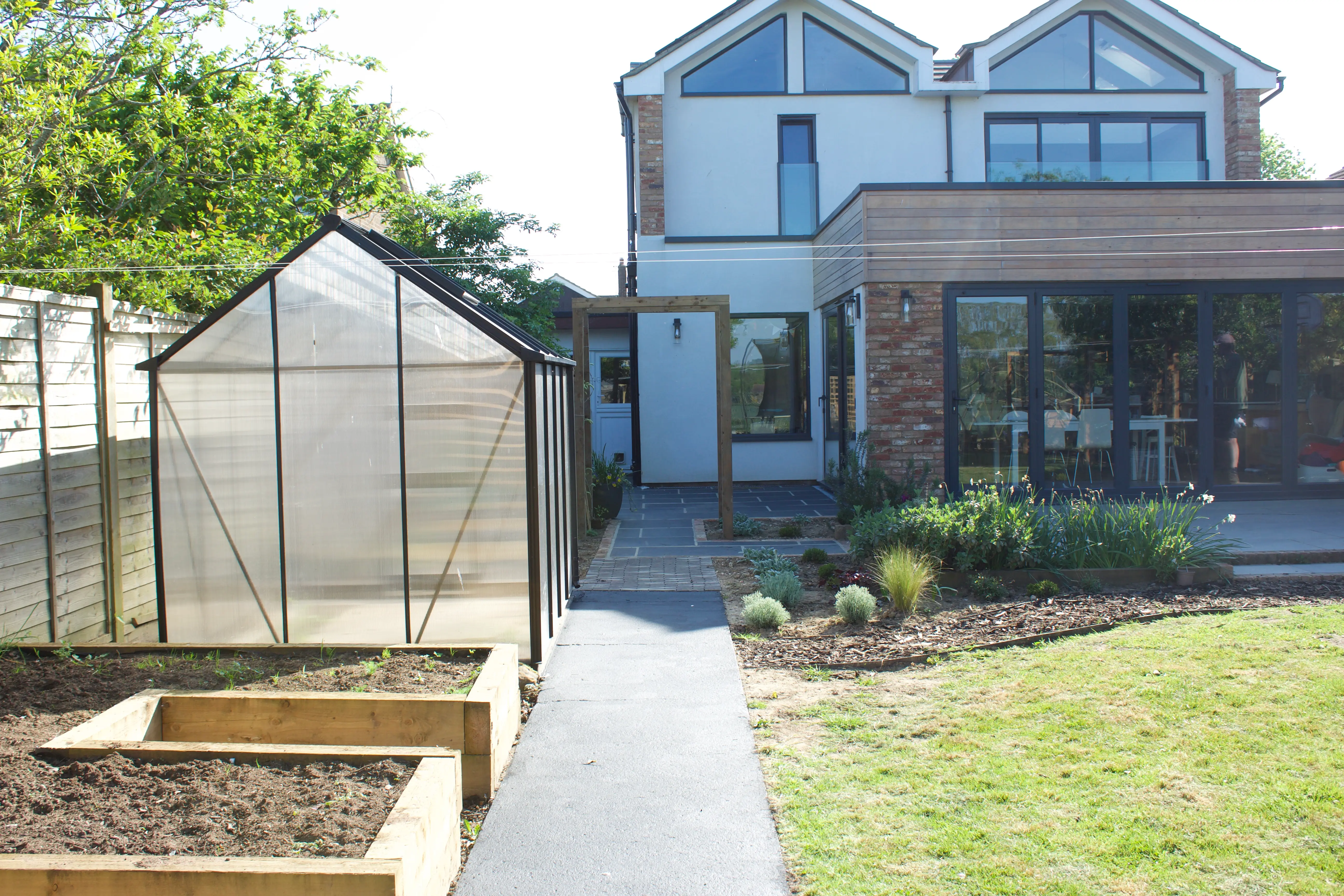 A backyard scene featuring raised garden beds, a greenhouse, and a two-story house in the background.