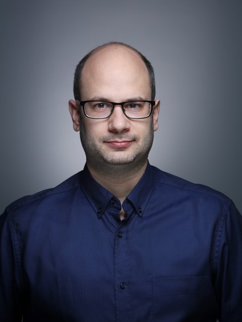 Young man in a dark suit jacket and cream sweater vest over a blue shirt with a patterned tie. Professional and confident expression, neutral background.