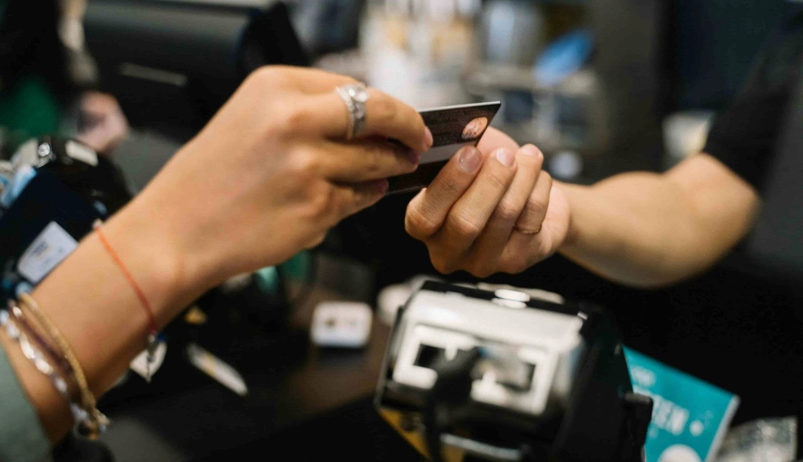 Mão de cliente entregando cartão de crédito para atendente em um balcão com terminal de pagamento ao fundo.