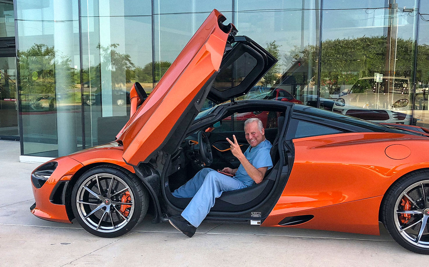 Dr. Billy (Dr. WIlliam Cohn) in blue scrubs sitting in an orange McLaren sports car with its scissor door open, smiling and flashing a peace sign in front of a glass-walled dealership.