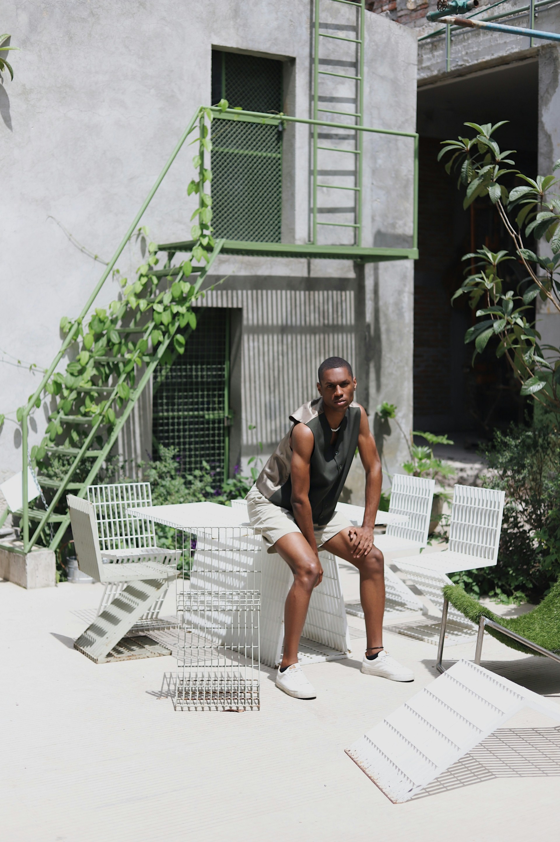 A man in an olive green vest and tan shorts sits on a white table in an industrial outdoor courtyard. Behind him, a green metal staircase with climbing vines leans against a grey concrete building.