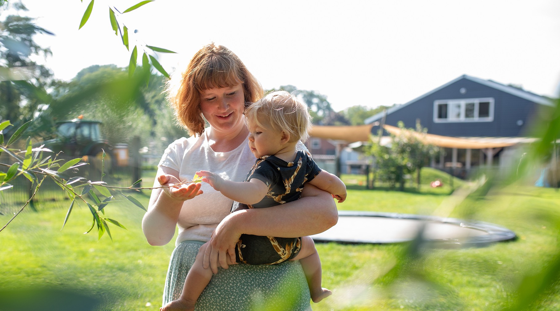 Een jonge jongen met blond haar en een groen T-shirt schommelt lachend op een autoband aan een rood touw in een groene, boomrijke speeltuin.
