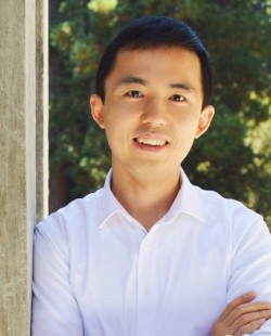 A smiling person wearing a white shirt, leaning against a wooden post with greenery in the background.