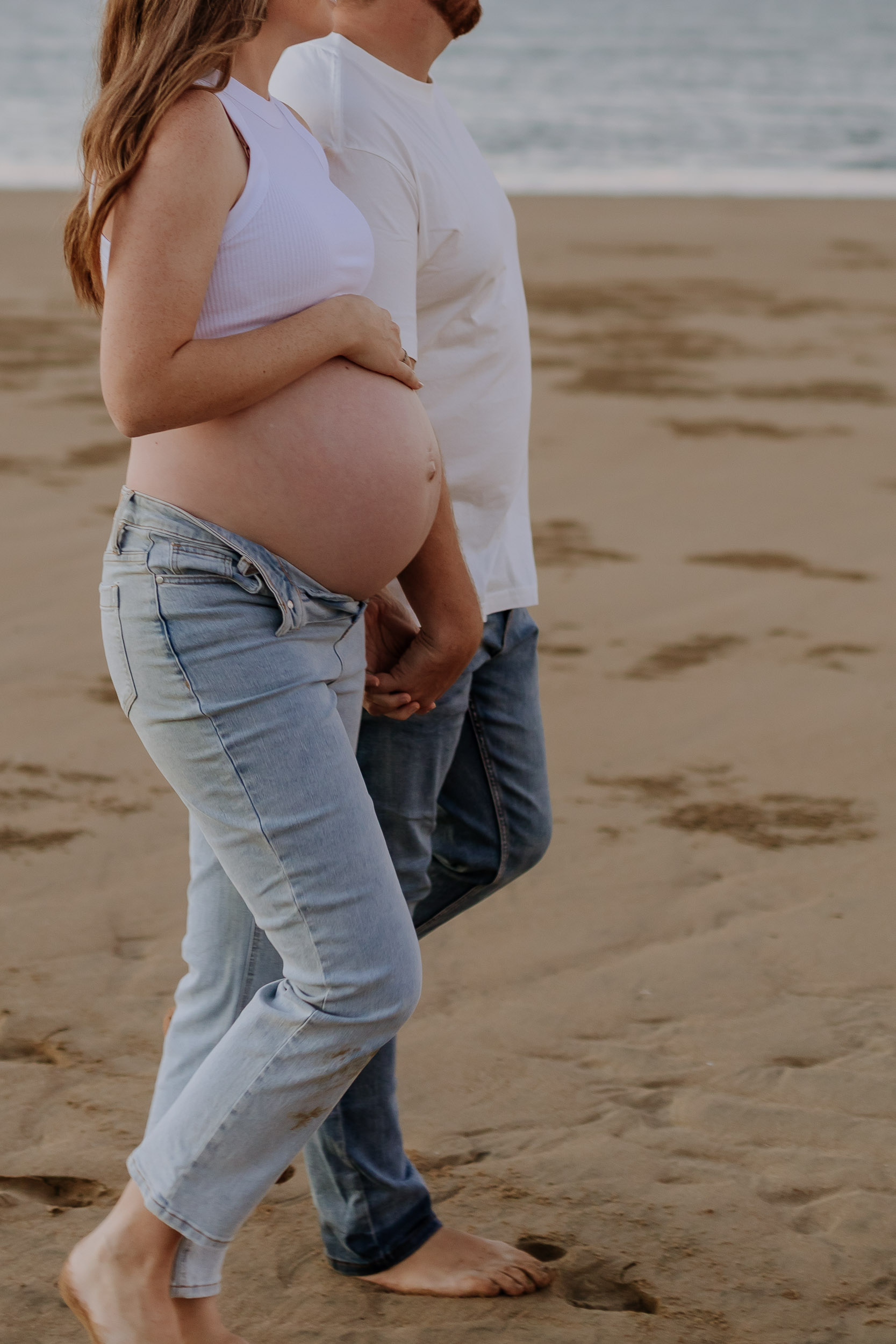 Pregnant couple walking along the beach holding hands with baby bump out