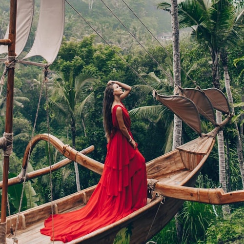 A woman in a red flowing dress stands on a wooden boat with lush greenery and palm trees in the background.