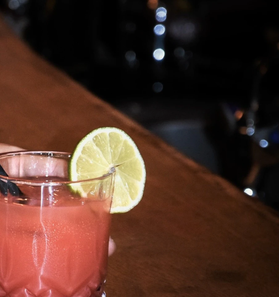 Rosafarbener Cocktail in einem Glas mit Zitronenscheibe am Rand, auf einer Bar.