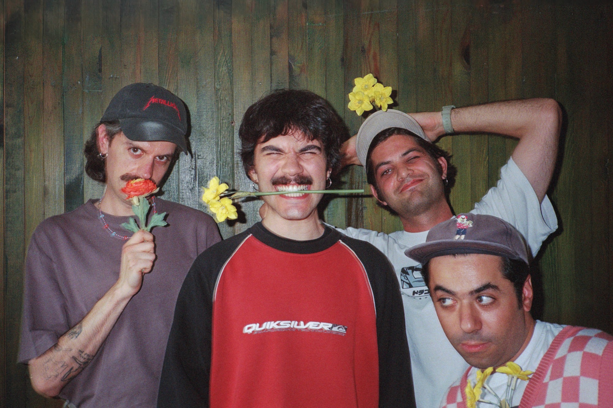 Four individuals pose playfully against a wooden backdrop, each holding colorful flowers, including a red poppy and yellow daffodils.