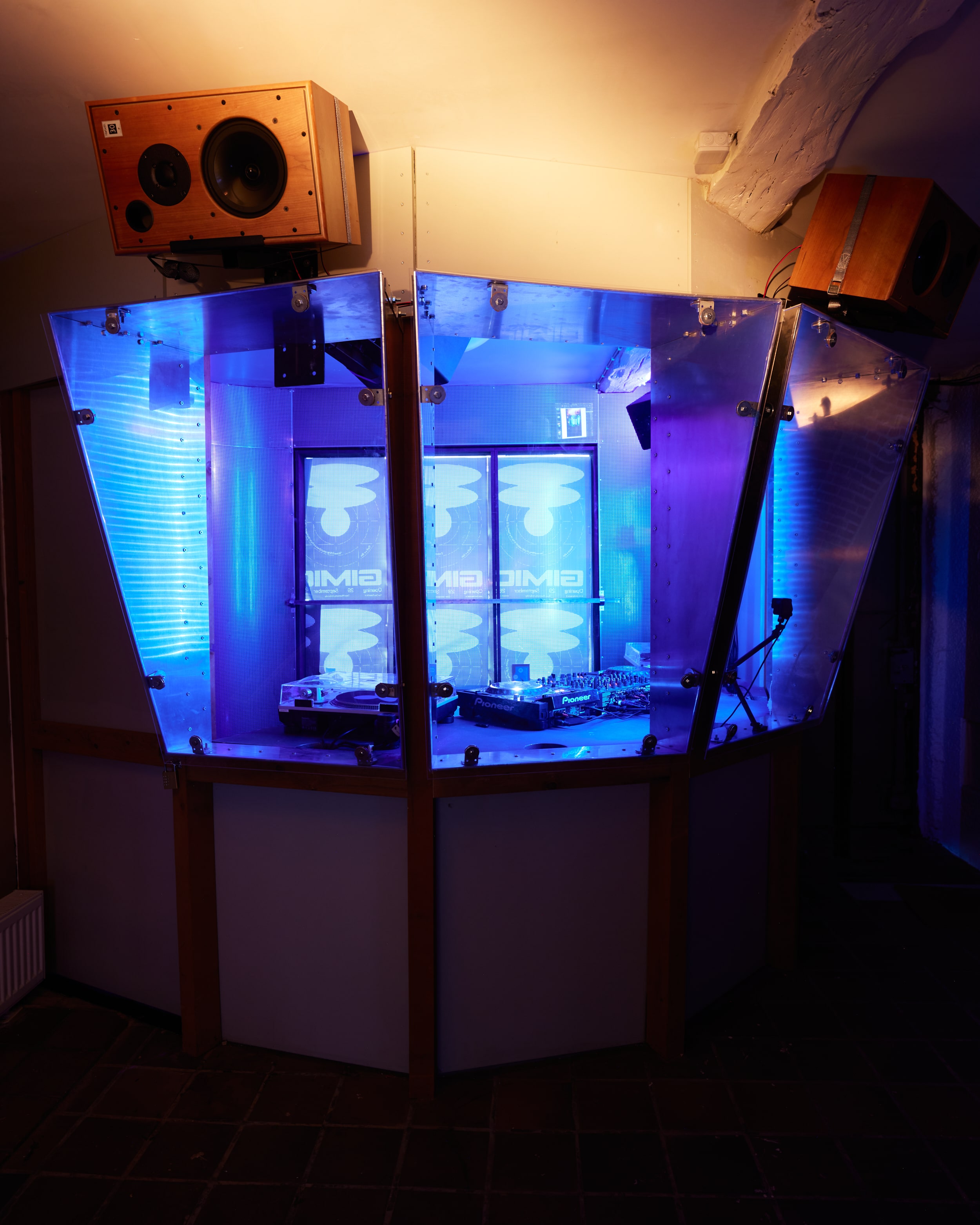 Blue-lit GIMIC DJ booth in a metal and plexiglass enclosure with wooden speakers mounted above