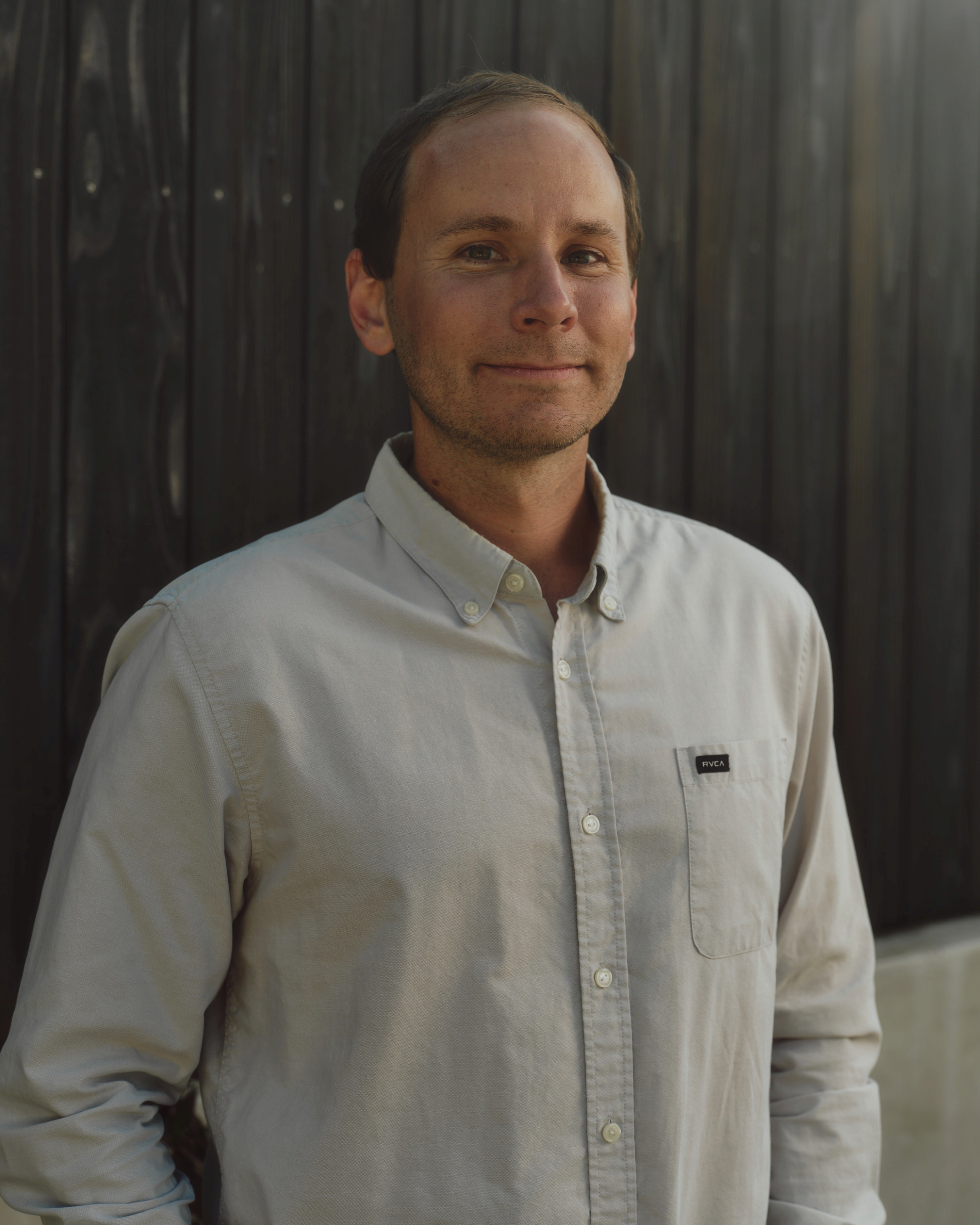 A portrait of a man with short brown hair, wearing a light-grey button-down shirt, standing in front of a dark vertical wood slat wall.