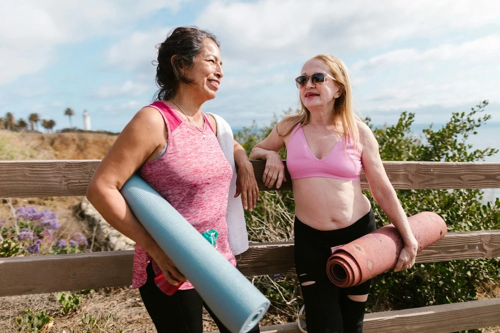 Two relaxed looking women smile and chat outdoors near a wooden fence with ocean views, each holding a yoga mat.
