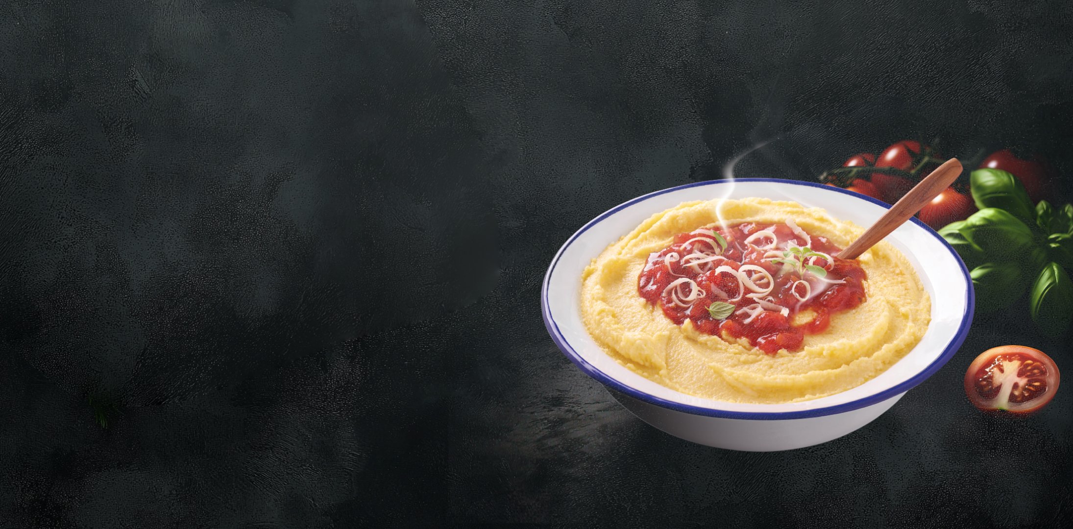 A steaming bowl of creamy polenta topped with a vibrant tomato sauce and garnished with sliced onions and herbs, sits alongside fresh basil and tomatoes on a dark background.