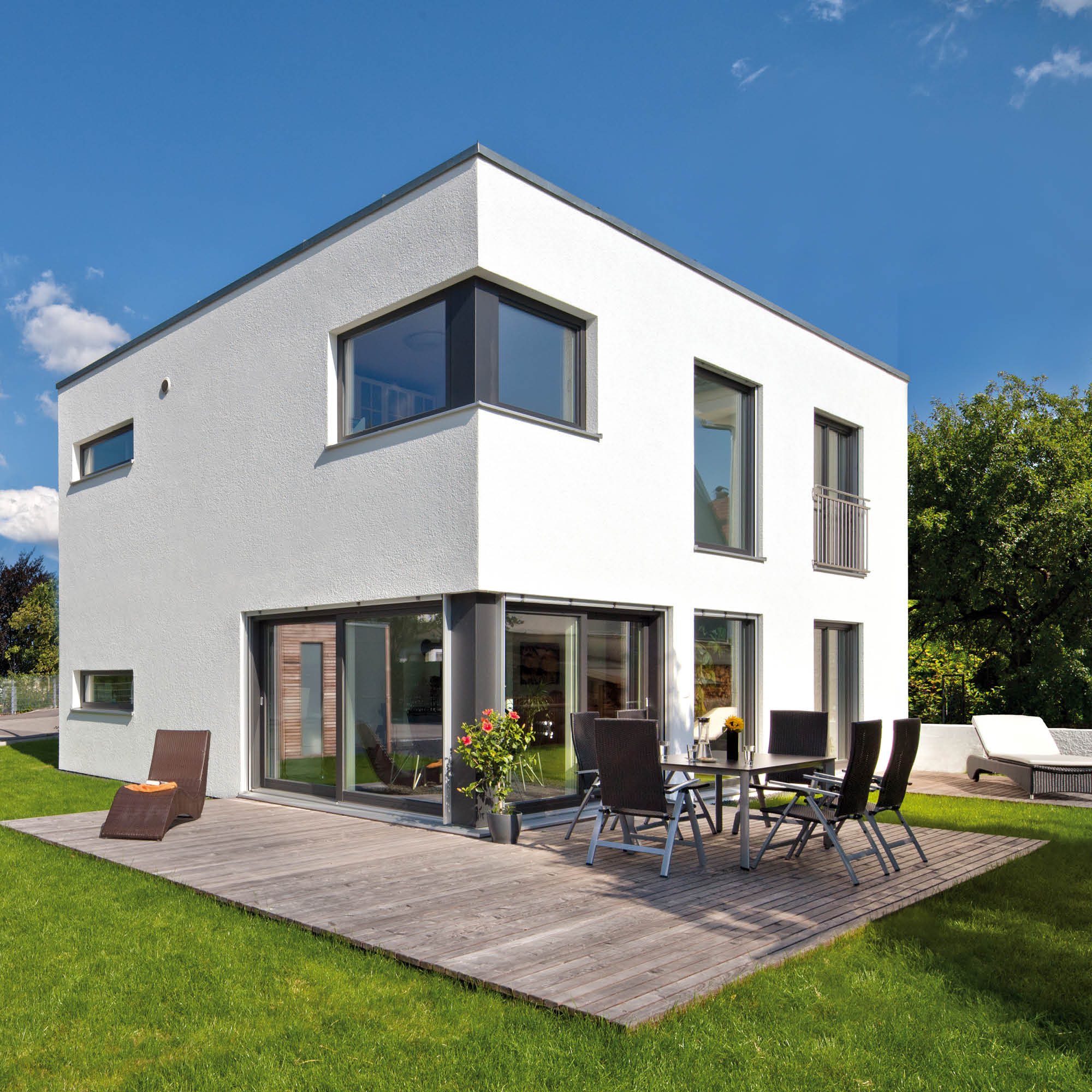 Moderner Neubau mit weißer Fassade, großer Holzterrasse, Gartenmöbeln und gepflegtem Rasen unter blauem Himmel.