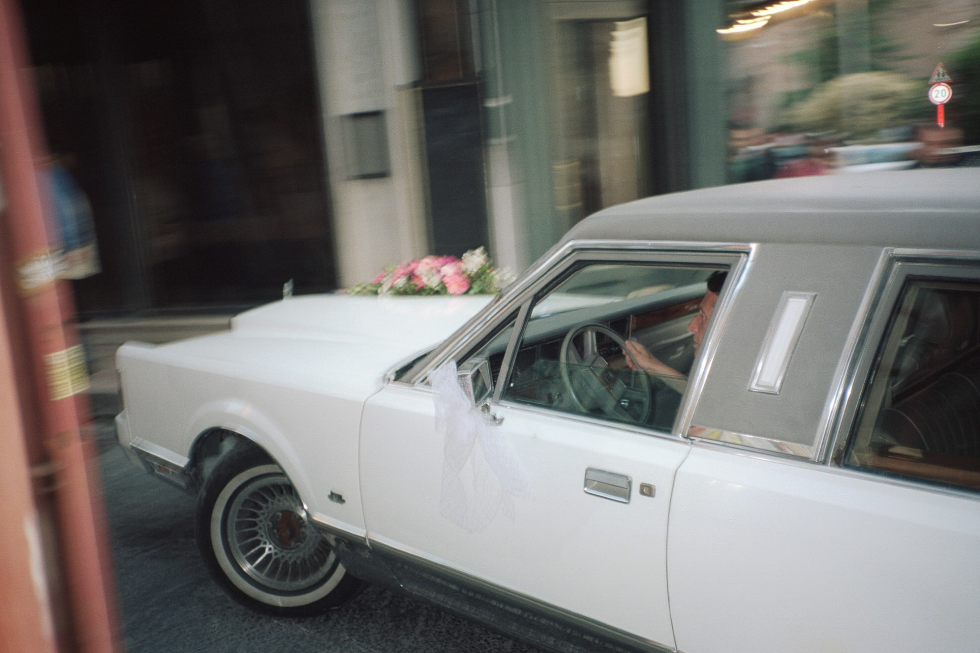 A white vintage car with a gray vinyl top, adorned with floral decorations, moves along an urban street, capturing the essence of a classic, elegant ride.