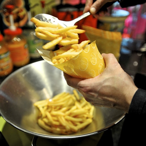 Valaki egy lyukacsos kanállal tölti meg a sárga tölcsért sült krumplival egy fém tálból. Különféle szószok vannak a háttérben.