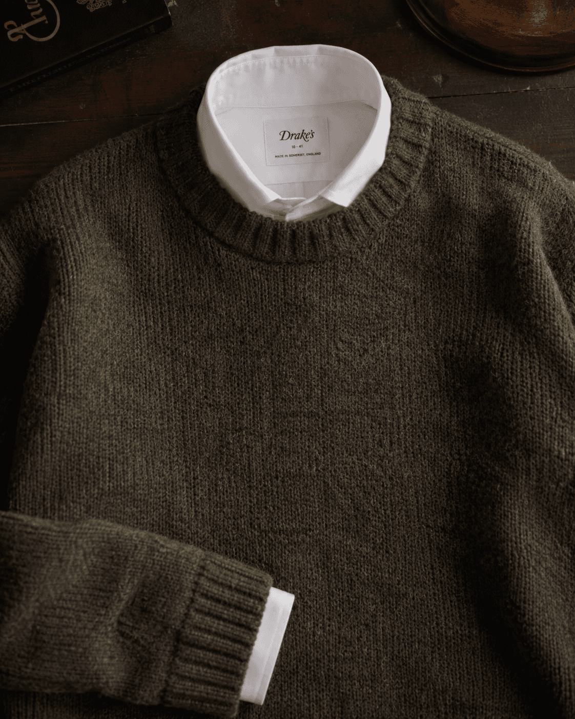 Alt Text:  Flat lay of a Drake's luxury menswear outfit featuring an olive green Shetland wool crewneck sweater layered over a white cotton dress shirt on a dark wooden table background.