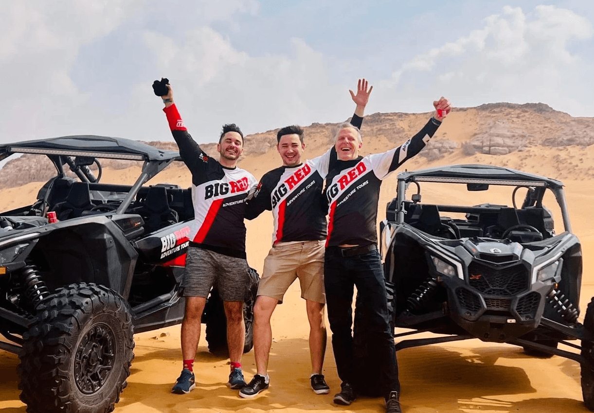 Three people celebrate in the desert by off-road vehicles, raising their arms with excitement against a rocky backdrop.