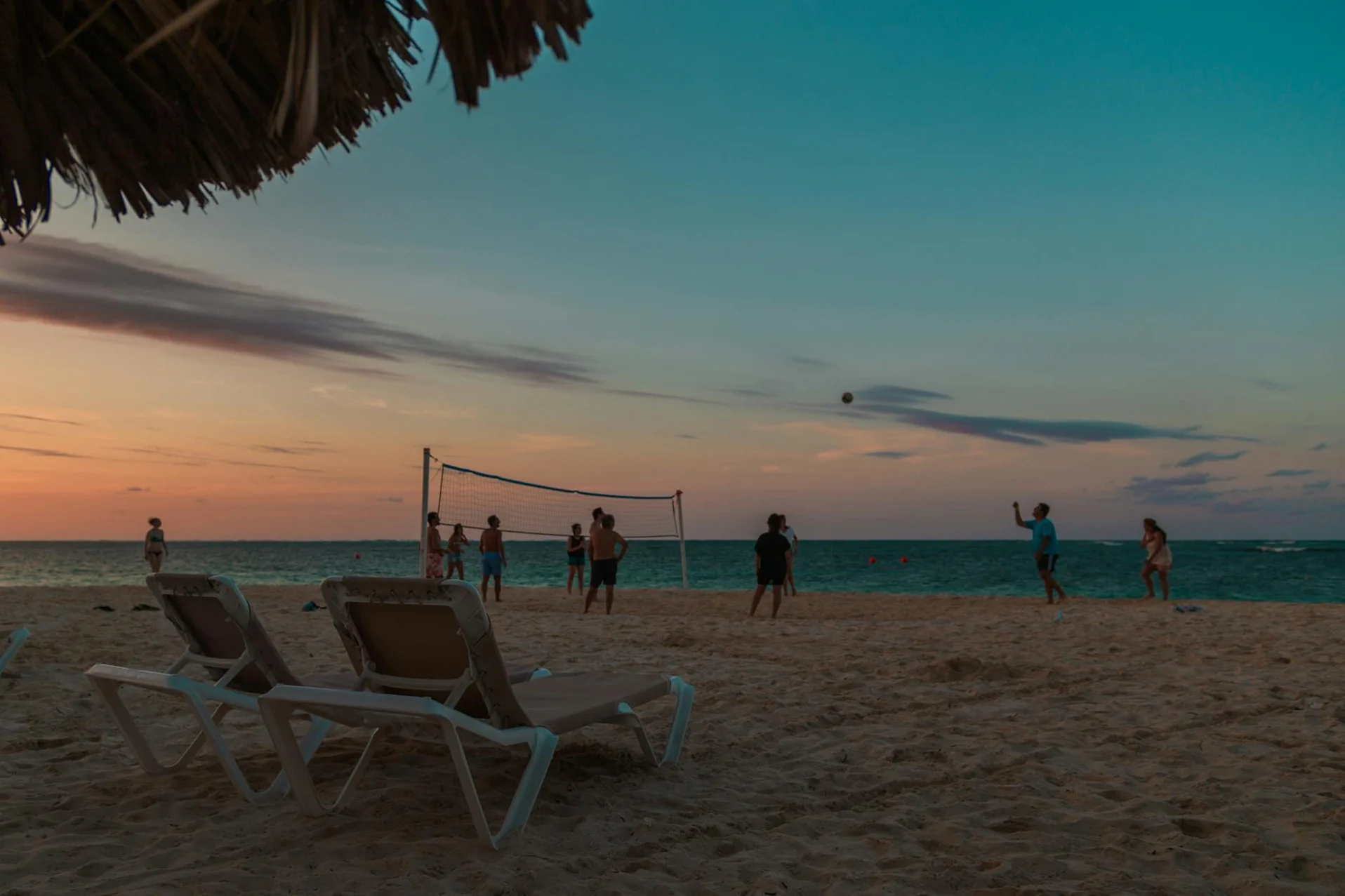 People playing beach volleyball at sunset near the ocean.