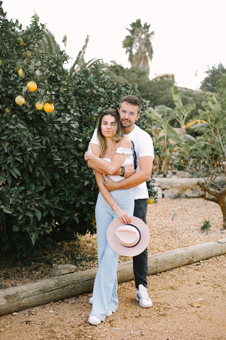 A woman with long blonde hair, in a brown top and jeans, walks through a sunlit forest, holding someone's hand. The scene is warm and inviting.