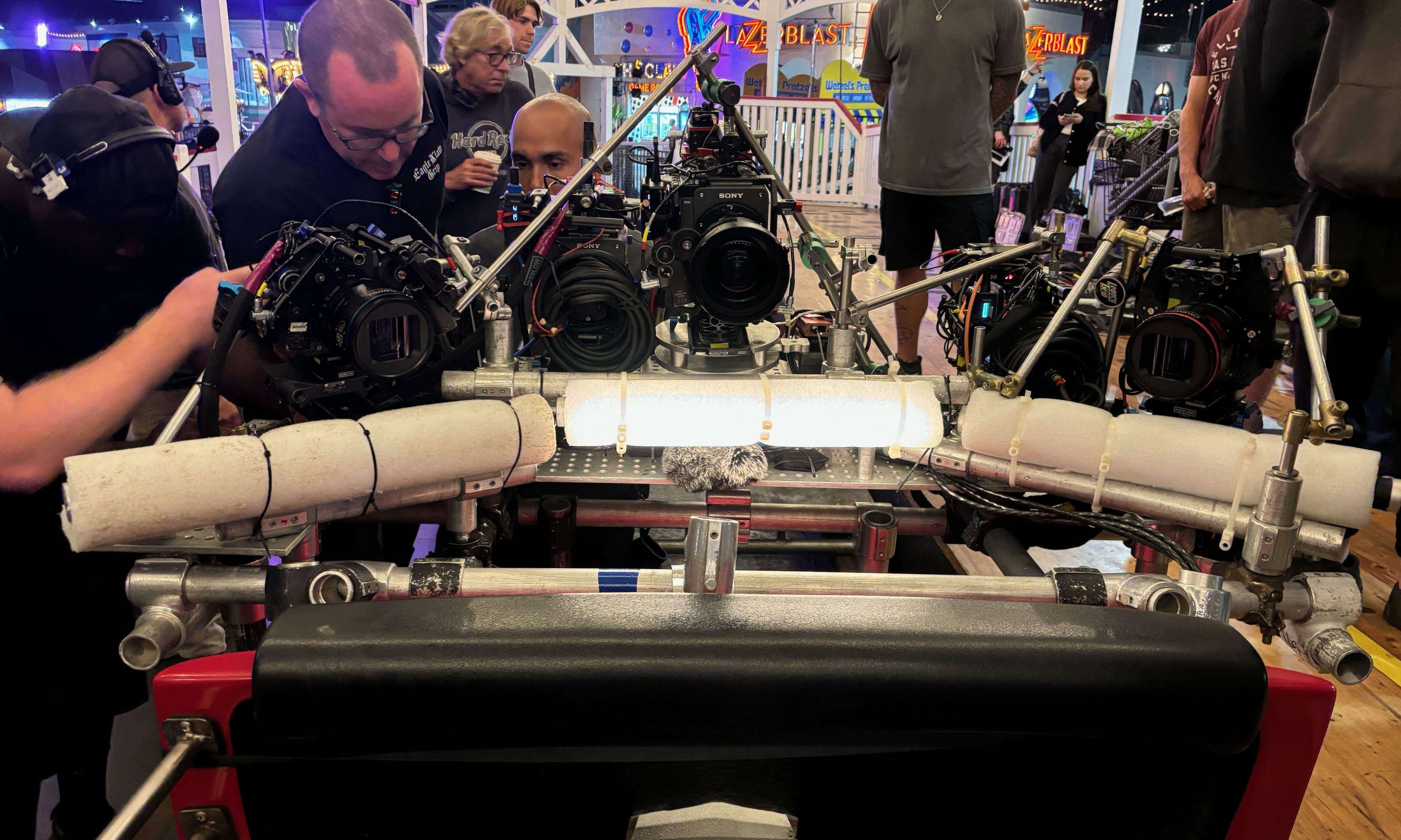 Three Sony cameras and an LED light panel mounted on a speedrail and cheeseplate rig on a roller coaster car at Belmont Park during a night shoot, with foam vibration padding visible and film crew surrounding the setup