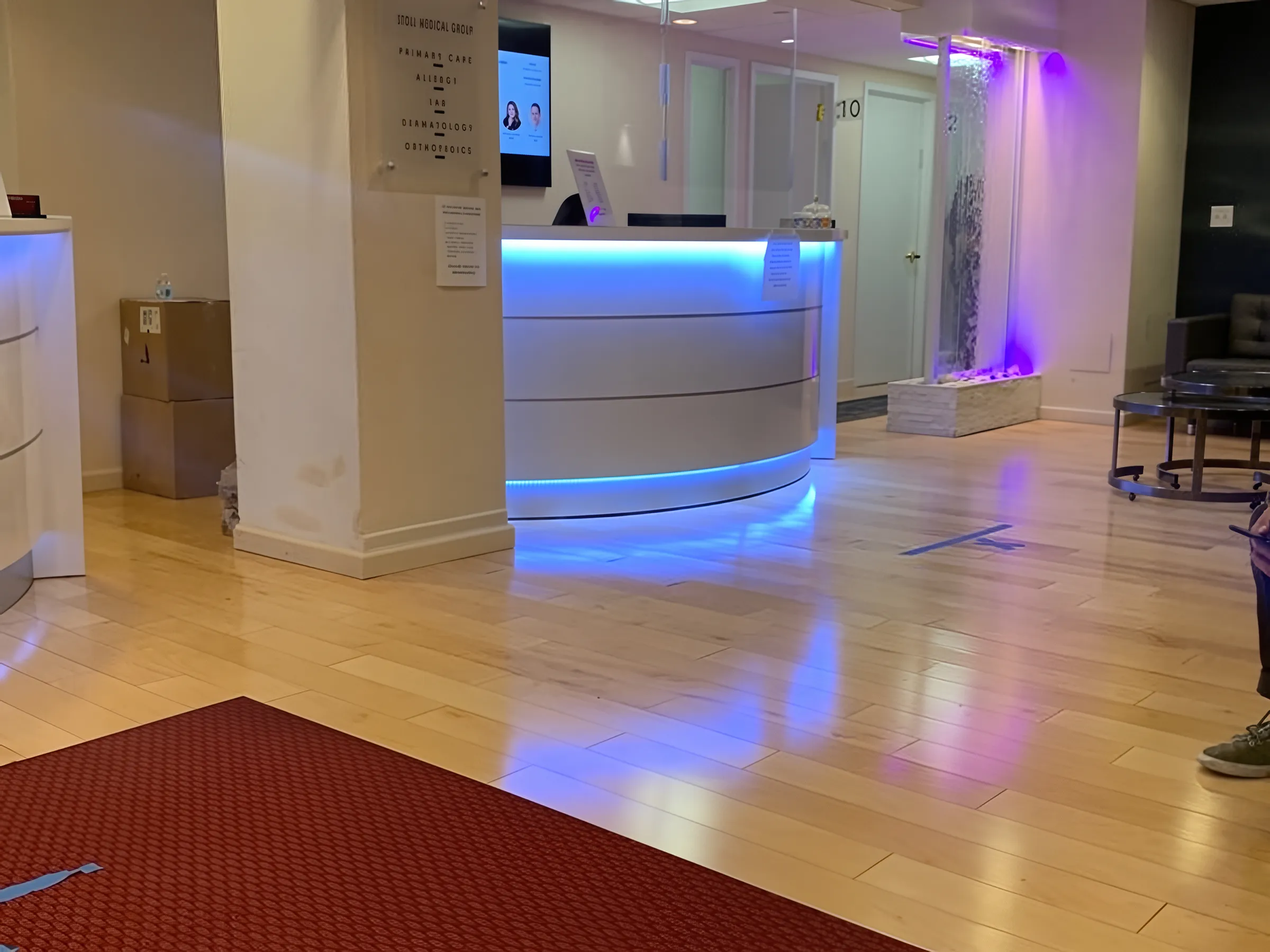 Hardwood flooring in a Stoll Medical Group open reception lobby with a curved LED-lit desk, water feature, and glowing accent lighting.
