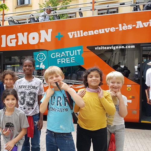 Skupina usmívajících se dětí pózuje před oranžovým vyhlídkovým autobusem, na kterém je napsáno "Gratuit Free" a "Avignon".