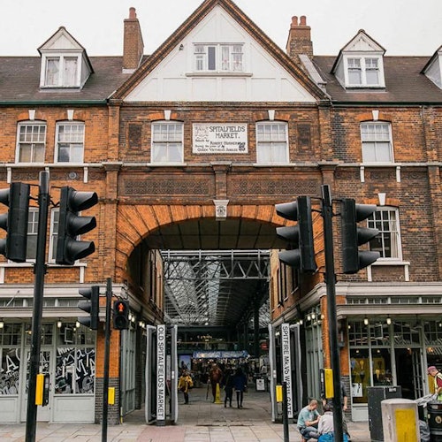 Isang pasukan sa Spitalfields Market sa ilalim ng gusaling gawa sa ladrilyo kung saan may mga taong naglalakad sa loob, napapalibutan ng mga ilaw trapo at mga tindahan.