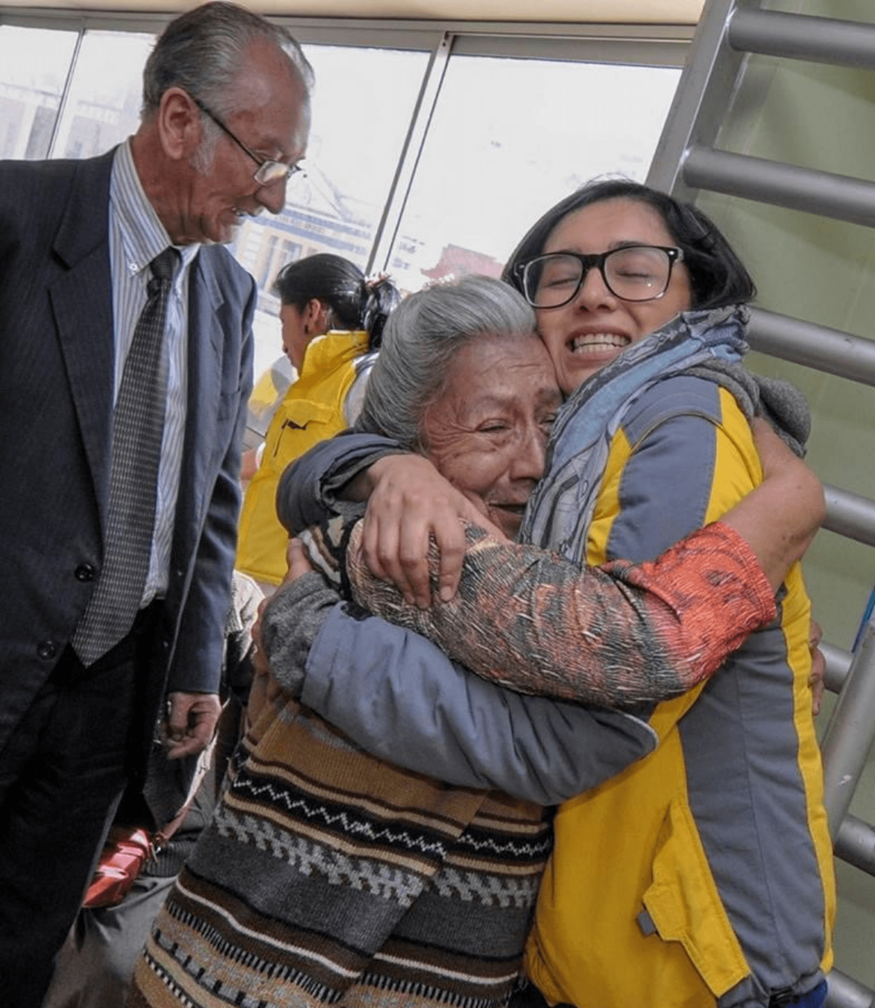 Daniela Jines warmly embraces an elderly woman while community members gather at a social support event.