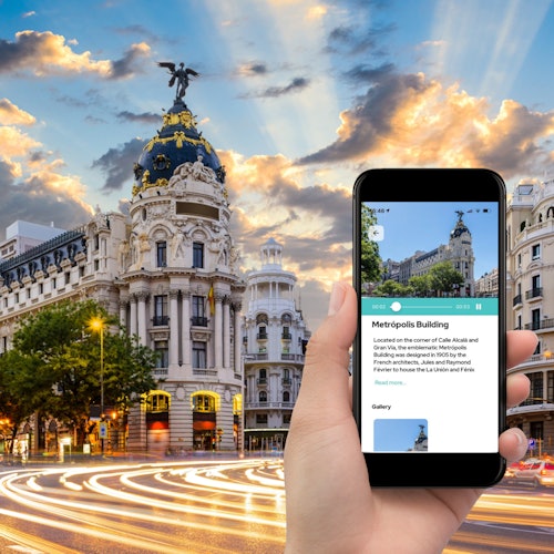 Ręka trzyma smartfon wyświetlający informacje o budynku Metropolis, z rzeczywistym budynkiem i tętniącą życiem scenerią miasta w tle.
