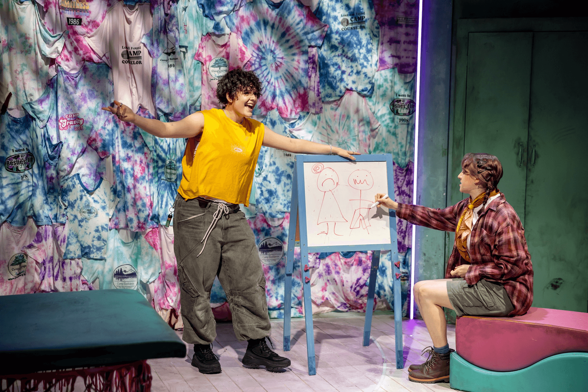A performer in a yellow sleeveless top gestures enthusiastically beside a small easel with a simple red marker drawing, while another performer sits beside them pointing at the sketch. The backdrop is covered in colourful tie-dyed camp T-shirts.