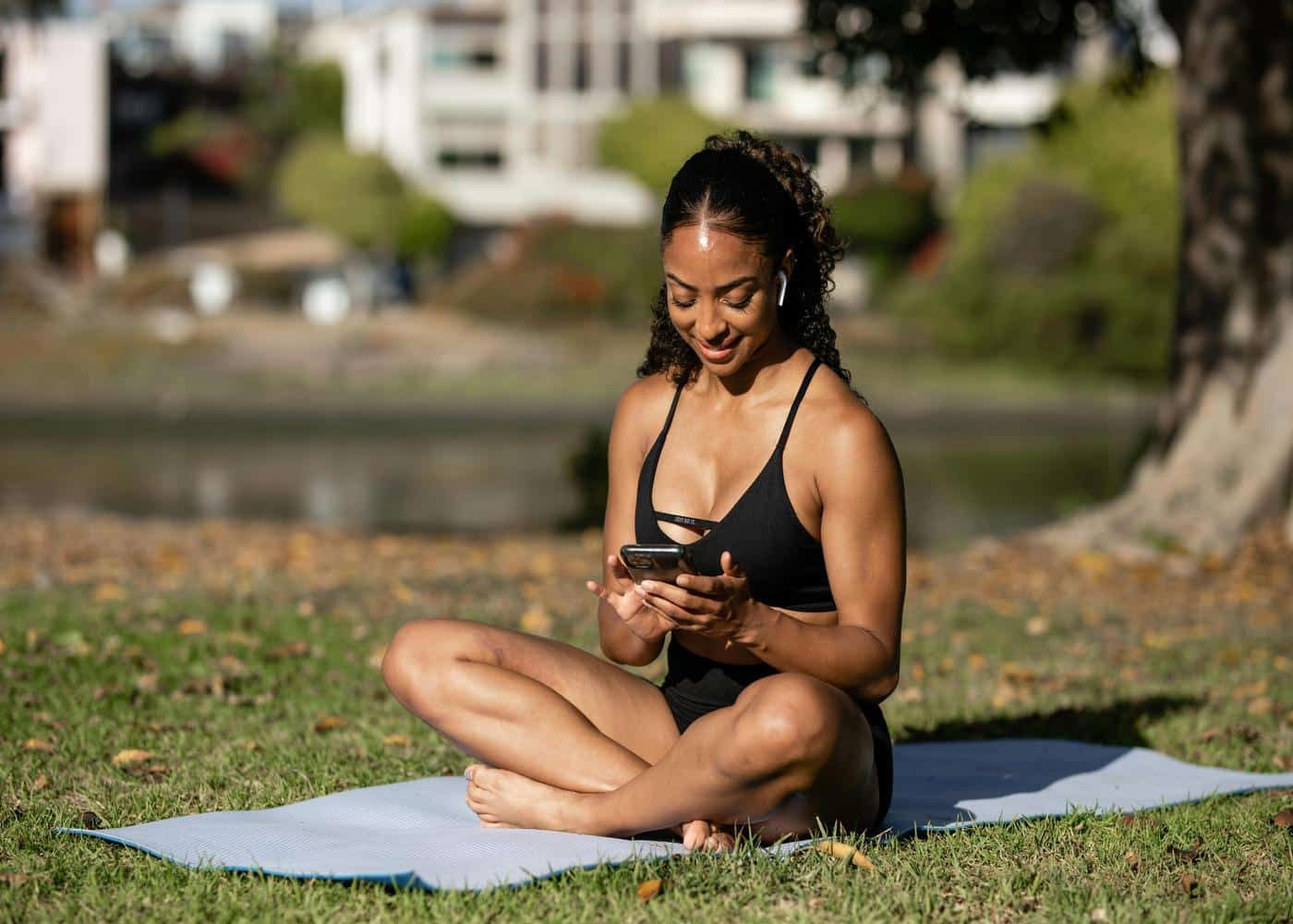Smiling woman sitting on yoga mat outdoors looking at smartphone