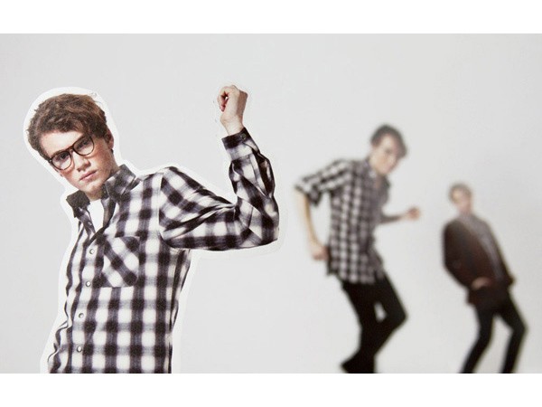 The image shows three men against a plain white background. The man in the foreground is wearing a black and white checkered shirt and glasses, and is flexing his bicep. His face is slightly turned toward the viewer. To his right, another man in a similar checkered shirt is mid-stride, appearing to be walking. The third man, on the far right, is dressed in a dark suit and is also in motion. All three men are out of focus, creating a shallow depth of field.