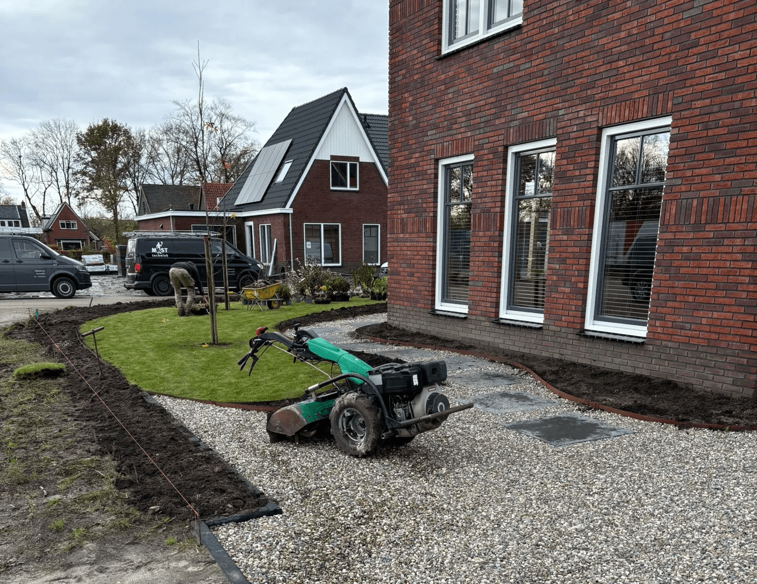 Grindpad met grote tegels langs bakstenen huis, omlijst door gras en verse aarde, hoveniers aan het werk bij nieuwbouw in Gorredijk.