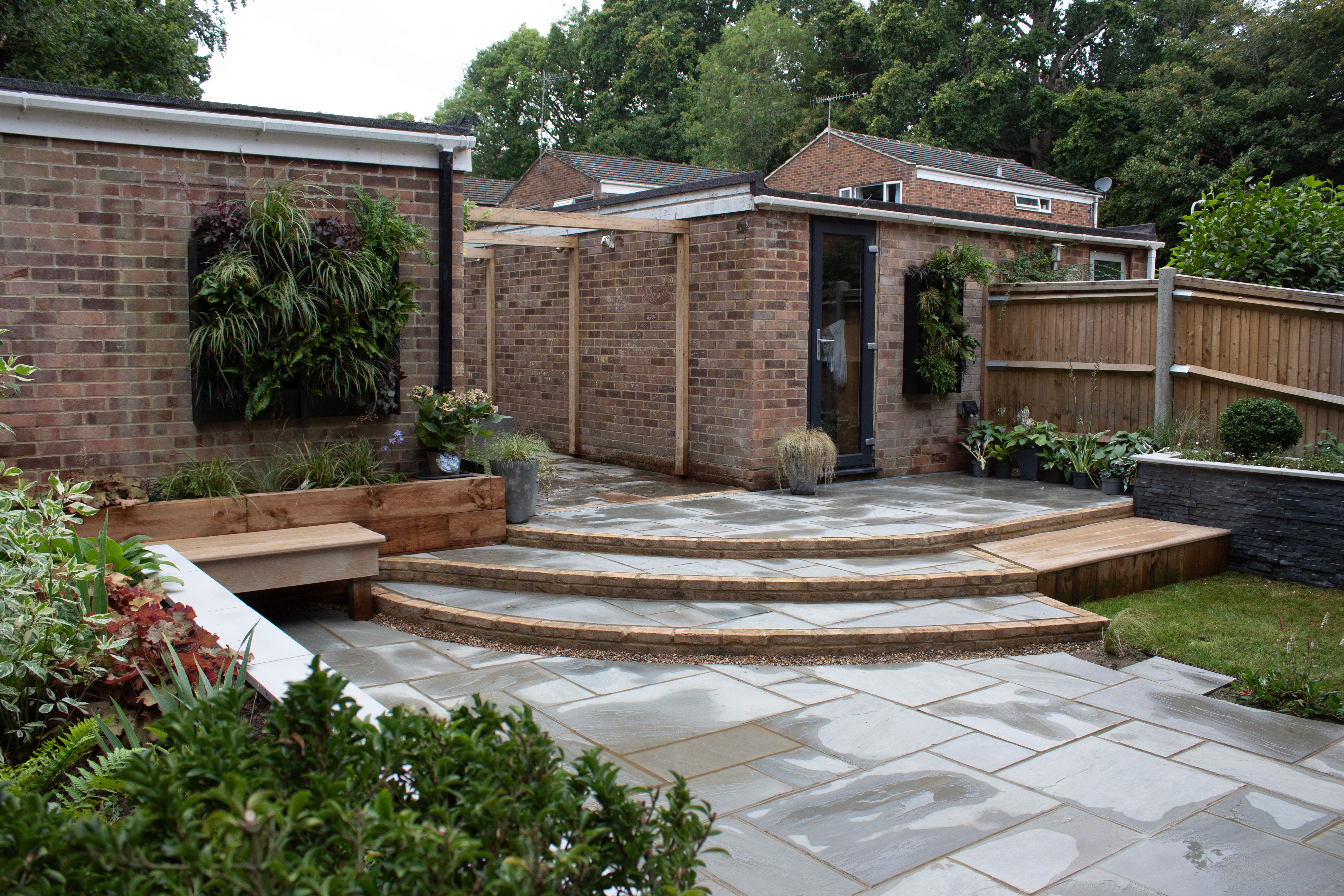 A modern home with brick walls and wooden deck steps leading to a backyard, surrounded by greenery.