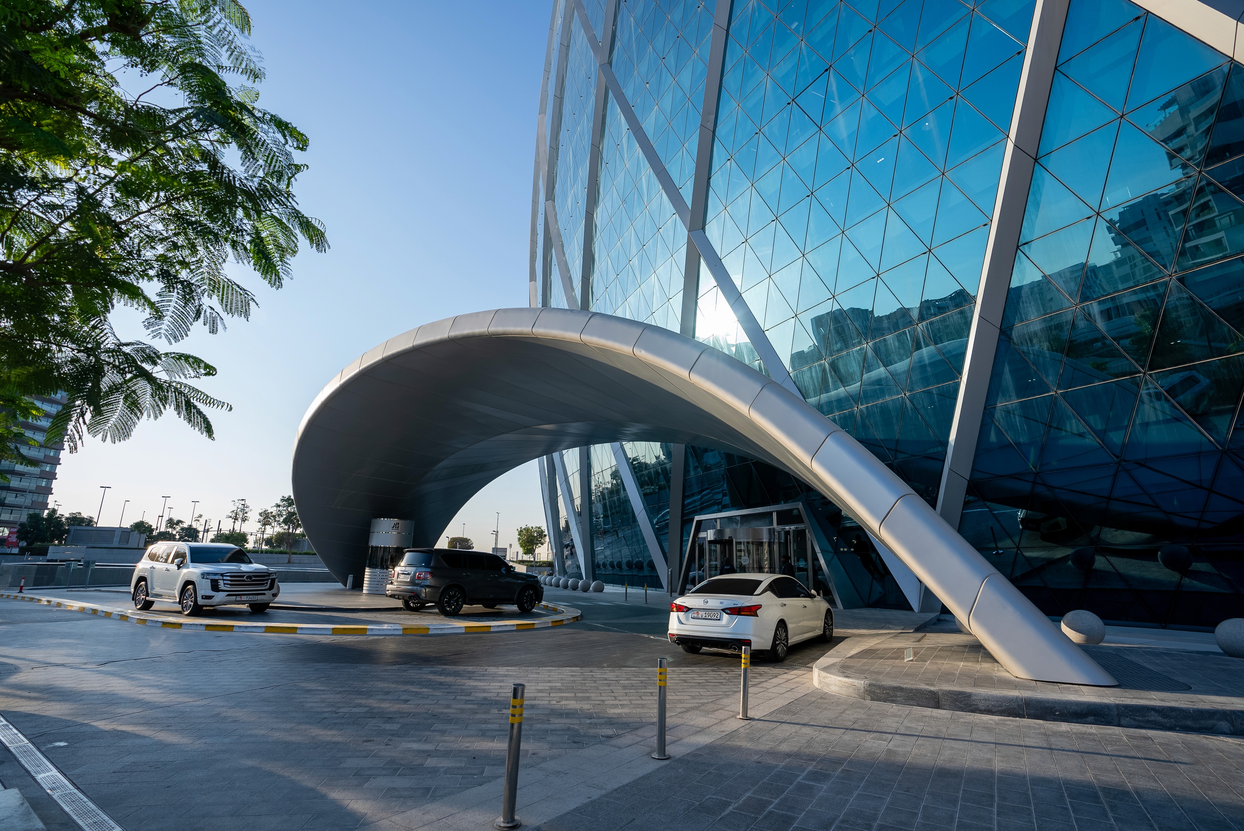 Exterior view of Office Square Aldar HQ, with a modern circular glass building and a curved silver canopy over the main entrance with parked cars.