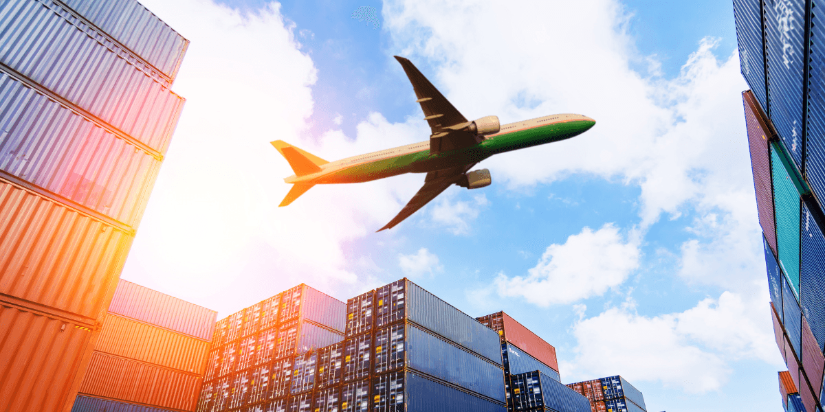 Airplane flying overhead above stacked shipping containers at cargo port under bright sky