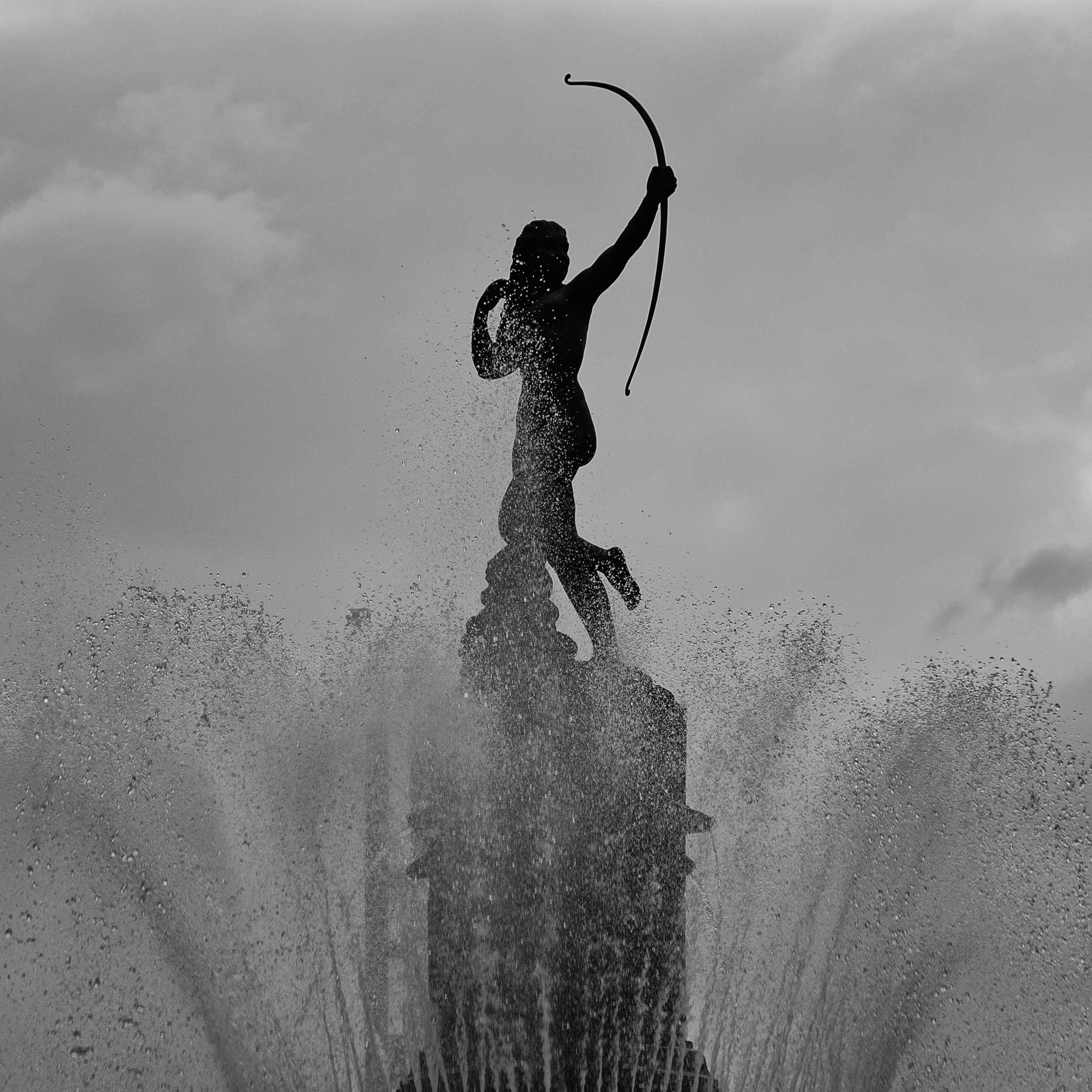 Black and white image of the Diana the Huntress statue in silhouette, bow raised, framed by rising fountain spray and a cloudy sky.