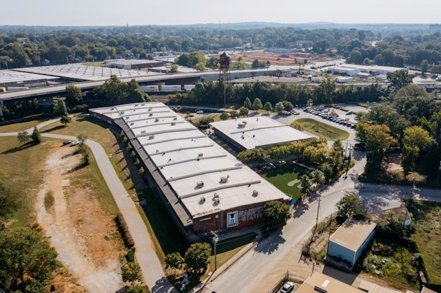 Aerial view of a manufacturing complex with multiple white-roofed warehouse buildings, surrounding vegetation, and an adjacent waterway