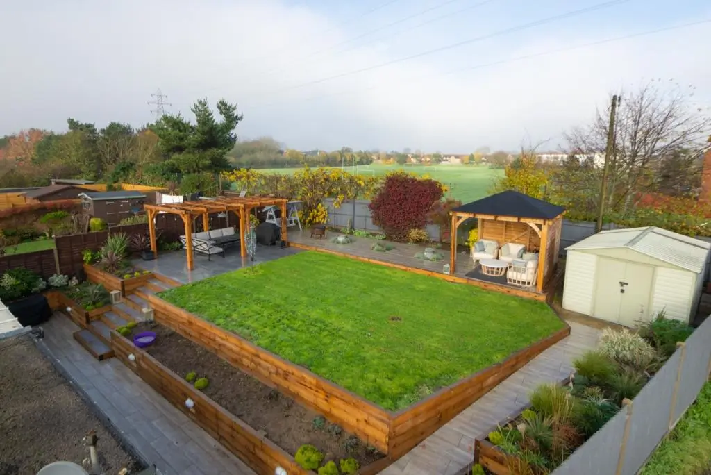 Aerial view of a landscaped garden featuring a green lawn, wooden fences, and garden structures in a serene setting.