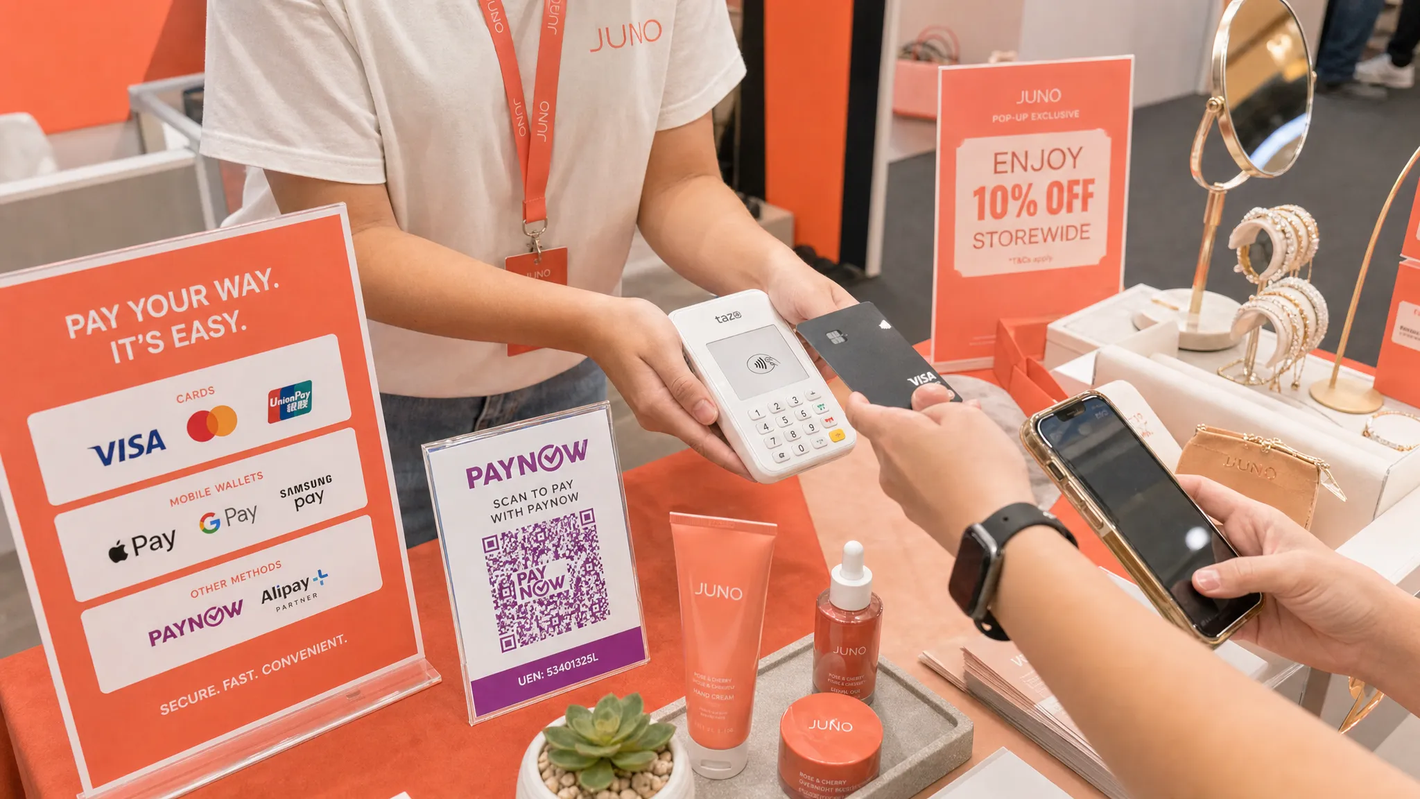 A compact Singapore pop-up booth checkout area with product displays, a visible PayNow QR stand, a smartphone being used for contactless card payment, and clear payment signage for cards and mobile wallets, shown from a slightly overhead angle with the checkout items arranged tightly on a small table.