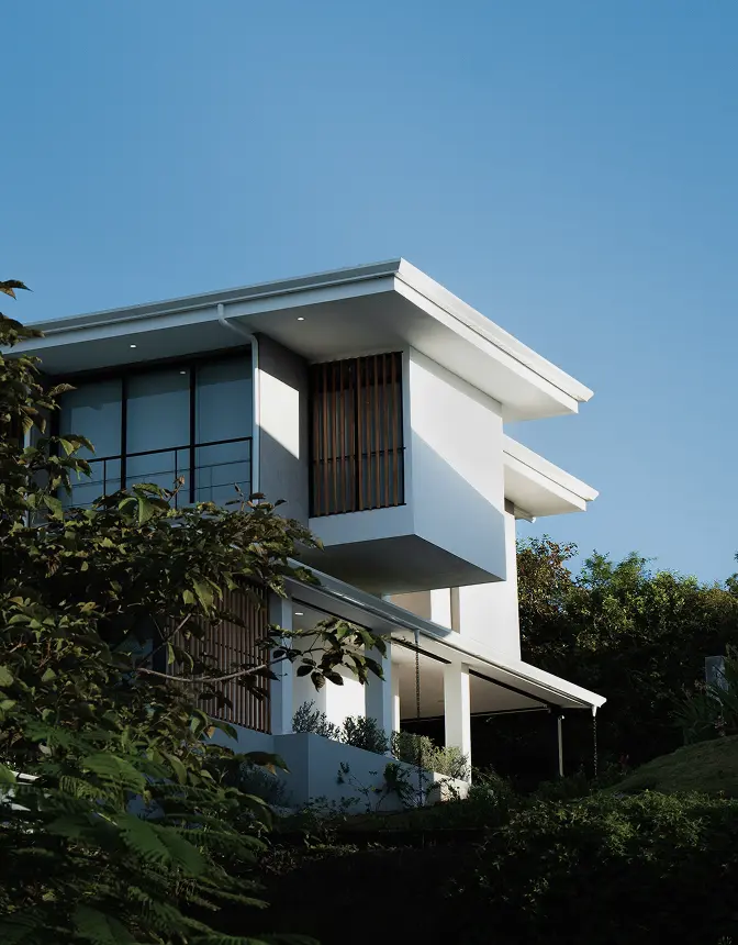 Modern white house with large windows and wooden accents, surrounded by greenery, set against a clear blue sky. The scene conveys tranquility.