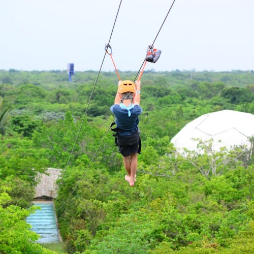 Person zip-lining over lush green forest, wearing a blue shirt, black shorts, and a yellow helmet. White dome structure in background.