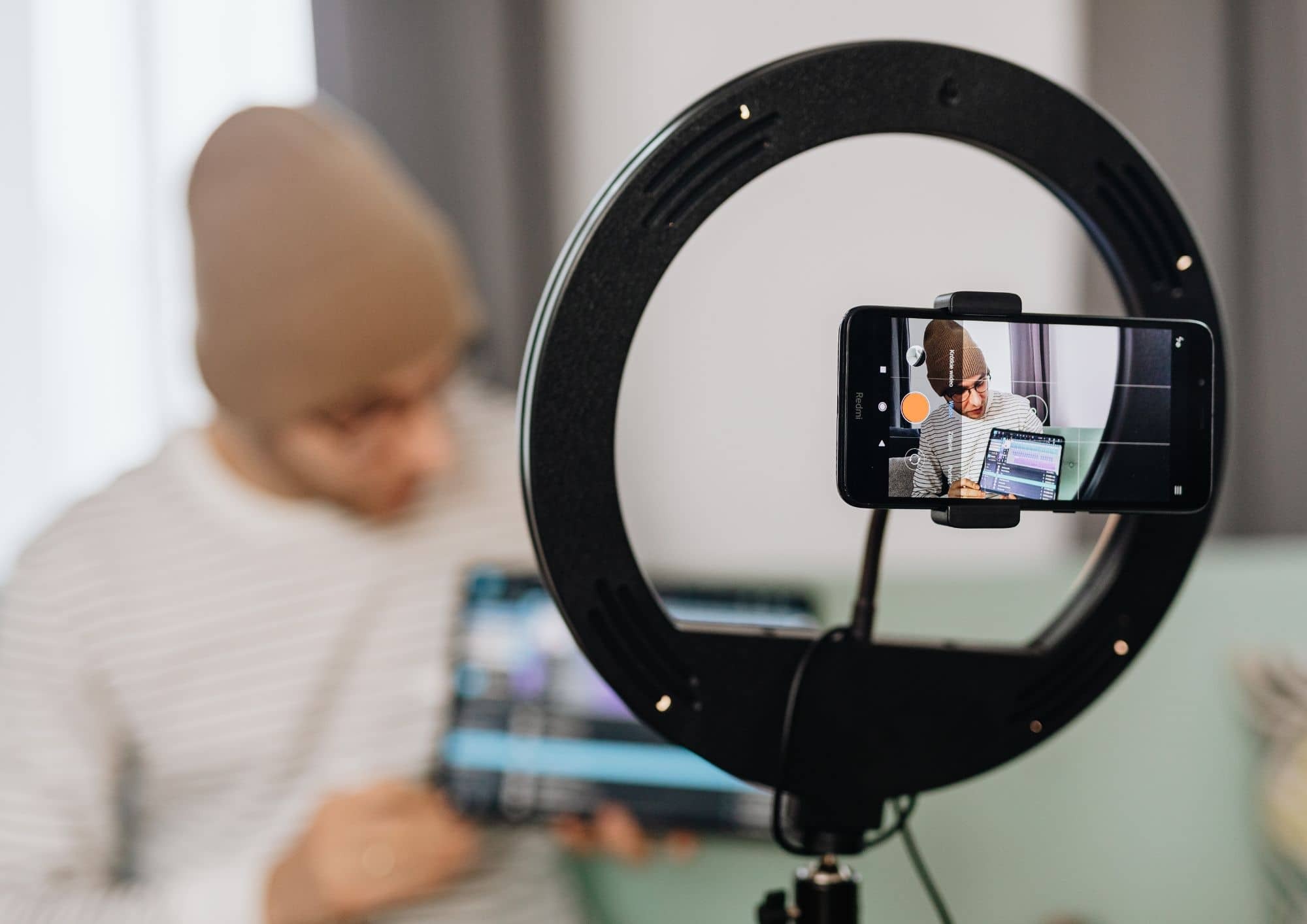 Pessoa grava um vídeo usando um smartphone preso a um ring light. Na tela do celular, ela aparece apresentando informações em frente a um computador, em um ambiente interno.