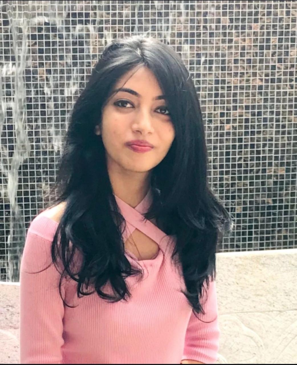 A smiling person with curly hair sits at a desk, looking at a laptop against a dark background.
