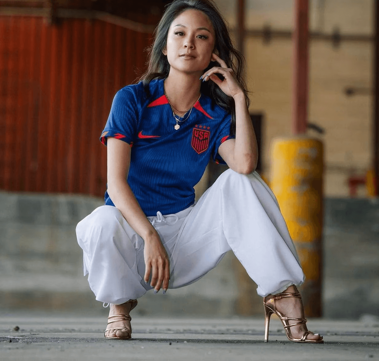 Meg Reyes in a blue USA soccer jersey, white pants, and gold high-heeled sandals crouches confidently indoors, looking at the camera. The background is industrial with concrete and red walls.