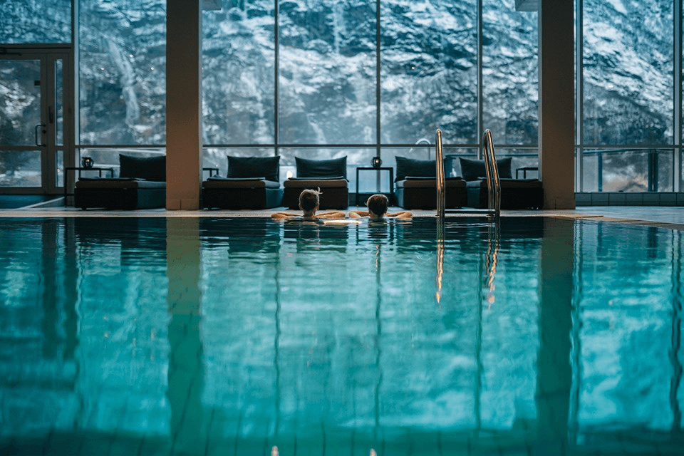 Two people relax in a luxurious indoor pool with large windows showcasing a scenic mountain view, capturing a serene and tranquil atmosphere.