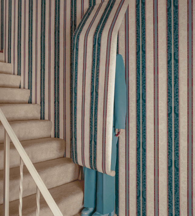 A person in blue is partially hidden behind a rolled piece of striped wallpaper that matches the striped wallpaper covering the walls and stairs in a surreal, visually intriguing manner.