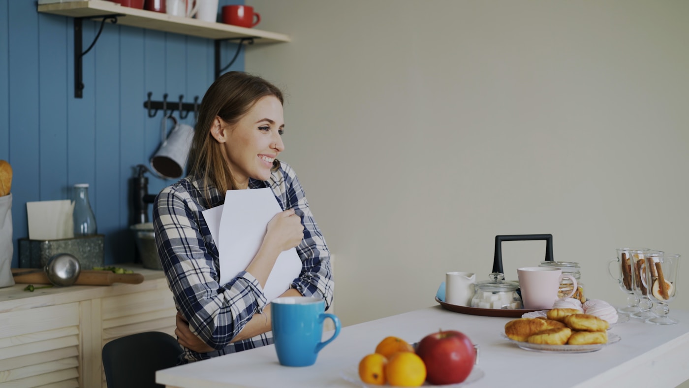 A woman smiling calmly at her kitchen table with fruit and a warm drink, suggesting relief and a comfortable relationship with food
