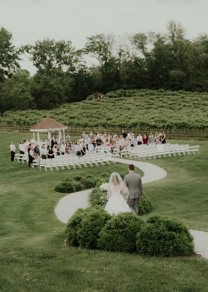 Aerial view of bride and groom walking down a winding garden path toward their guests and a gazebo at a vineyard wedding ceremony