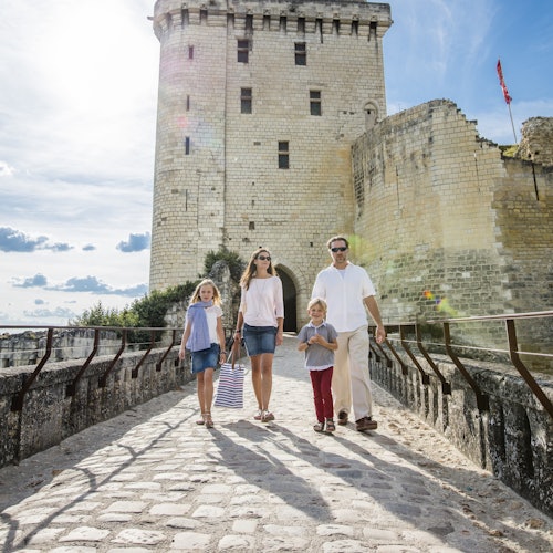 A family of four walks on a cobblestone path towards a historic stone castle under a sunny sky.