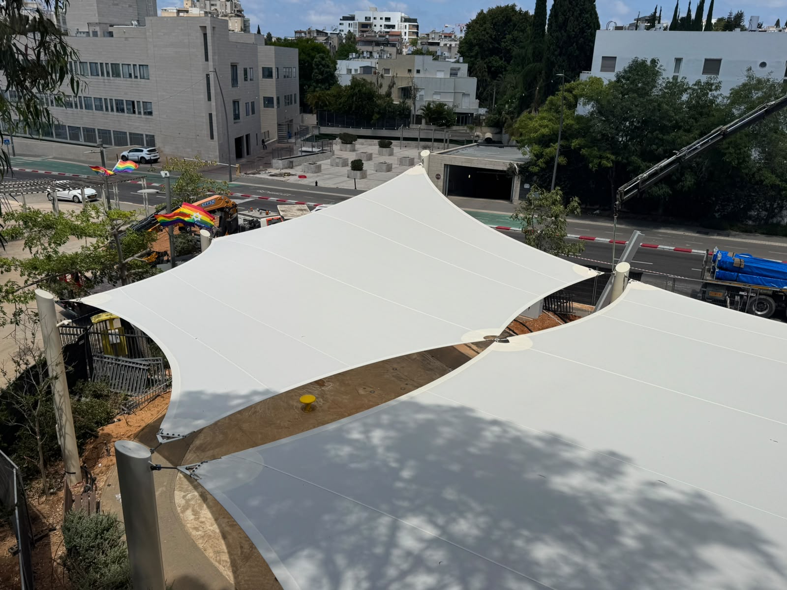 Two large white shade sails stretch over a paved area in an urban setting, with buildings and trees in the background.